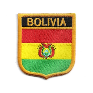 Puede incluir: Parche bordado con el diseño de la bandera boliviana. El parche tiene forma de escudo con un panel superior negro que dice "BOLIVIA" en letras doradas. El diseño de la bandera incluye franjas horizontales rojas, amarillas y verdes, con un escudo de armas en el centro.