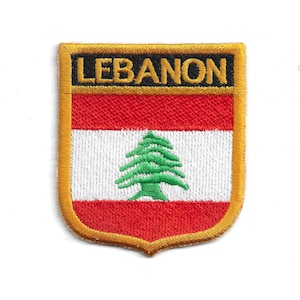 Może przedstawiać: Haftowana naszywka z flagą Libanu. Naszywka ma kształt tarczy ze złotą obwódką. Projekt flagi obejmuje czerwone i białe poziome paski, zielone drzewo cedrowe i napis "LEBANON" w kolorze czarnym.