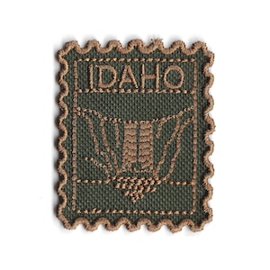 Peut inclure: Écusson brodé en forme de timbre-poste. Le patch est vert olive avec des coutures marron. Le mot "IDAHO" est cousu en haut. Le motif présente une illustration marron d'un bâtiment et un motif stylisé.