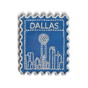 May include: A blue embroidered patch in the shape of a postage stamp. The word "DALLAS" is at the top, with a white outline of the Dallas skyline. The patch has a scalloped edge.