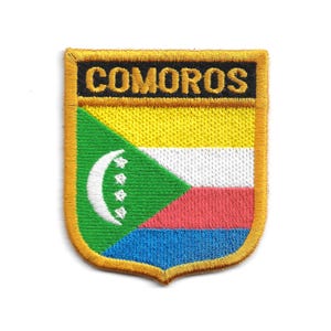 May include: Embroidered patch featuring the word "COMOROS" in gold lettering above a shield design. The shield displays the Comoros flag with green, yellow, white, red, and blue sections, and a crescent moon with four stars.