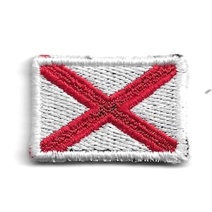 Könnte beinhalten: Ein weißer Stoff-Patch mit einem roten gestickten Kreuz. Der Patch hat einen schwarzen Rand.