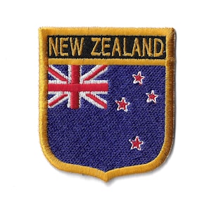 May include: A blue and white embroidered patch with a gold border. The patch features the New Zealand flag with the Union Jack in the top left corner and four red stars in the bottom right corner. The text "NEW ZEALAND" is embroidered in black on a gold background at the top of the patch.