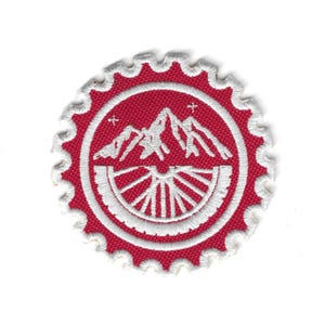 May include: Embroidered patch with a red background and a white mountain and wheel design. The patch has a scalloped edge and is circular in shape. The design includes a mountain range and a stylized wheel.