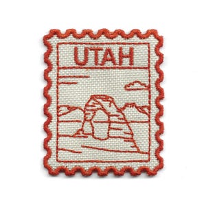 Puede incluir: Parche bordado con forma de sello postal, con la palabra "UTAH" en la parte superior. El diseño incluye un arco y una cordillera en el centro, con un borde rojo. El parche está sobre un fondo blanco.