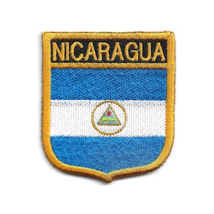 Può includere: Toppa ricamata con la bandiera del Nicaragua. Il design presenta la scritta "NICARAGUA" in lettere dorate, sopra strisce orizzontali blu e bianche. Un emblema circolare con una montagna e un triangolo è centrato.