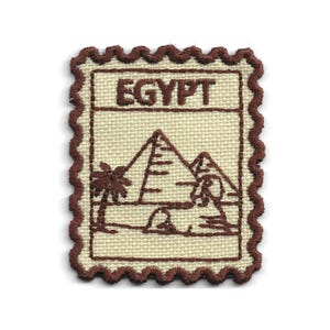 May include: A rectangular embroidered patch with a scalloped edge, featuring a cream-coloured fabric base and dark brown thread. The design depicts the word EGYPT above a scene with pyramids, a palm tree, and a sphinx, all outlined in brown.