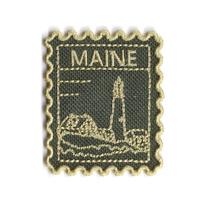 Puede incluir: Parche bordado con forma de sello postal, con un faro y la palabra "MAINE". El parche tiene un fondo verde oscuro con bordado dorado y un borde festoneado.