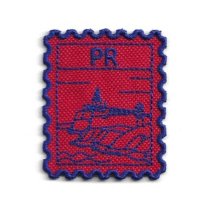 Può includere: Una toppa in tessuto rosso a forma di francobollo, con un bordo blu smerlato. Presenta le lettere "PR" e un'illustrazione blu di un edificio e onde.