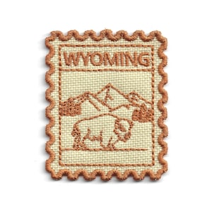 May include: Embroidered patch featuring a tan background with a brown border and design. The design includes the word "WYOMING" above a mountain range and a bison. The patch has a postage stamp design.