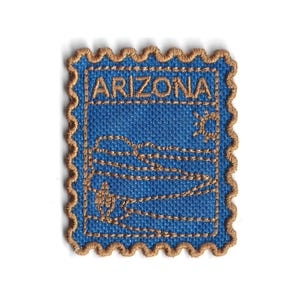 Puede incluir: Parche bordado con forma de sello postal. El parche presenta la palabra "ARIZONA" en hilo dorado sobre un fondo azul. El diseño incluye un sol, un paisaje desértico y un cactus. El parche tiene un borde decorativo festoneado.