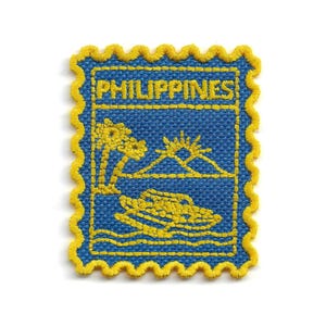 May include: A rectangular embroidered patch with a scalloped yellow border on a blue background. The text PHILIPPINES is stitched in yellow at the top. Below, yellow embroidery depicts palm trees, a sun over a mountain, and a boat on waves.