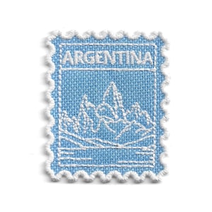 Puede incluir: Un parche bordado rectangular con forma de sello postal, con una base de tela azul claro y detalles en hilo blanco. El diseño presenta la palabra ARGENTINA en letras mayúsculas blancas sobre un paisaje de montaña bordado en blanco.
