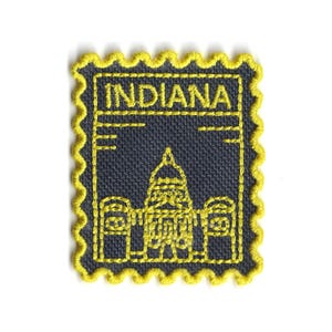 Peut inclure: Écusson brodé avec le mot "INDIANA" et une illustration d'un bâtiment, probablement un capitole d'État. L'écusson a un fond bleu foncé, des coutures jaunes et un bord festonné. Le design ressemble à un timbre-poste.
