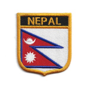 Puede incluir: Parche bordado con la palabra "NEPAL" en letras doradas sobre fondo negro. El parche en forma de escudo muestra la bandera de Nepal, con un campo rojo, borde azul y emblemas blancos. El parche está bordeado con hilo dorado.