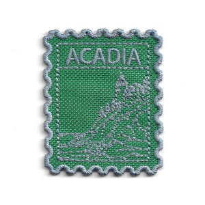 May include: Embroidered patch in the shape of a postage stamp. The patch features the word "ACADIA" in grey thread above a grey illustration on a green background. The border is a scalloped edge in grey.