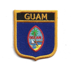 May include: Embroidered patch featuring the word "GUAM" in gold on a black banner. Below, a blue shield displays a Guam seal with a palm tree, sailboat, and the word "GUAM". The patch has a gold border.