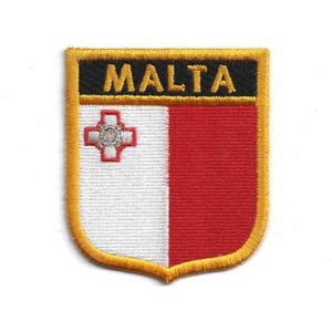 May include: Embroidered patch featuring the flag of Malta. The patch is shield-shaped with a gold border. The flag design includes a white and red vertical split, with the word "MALTA" in gold letters on a black banner at the top.