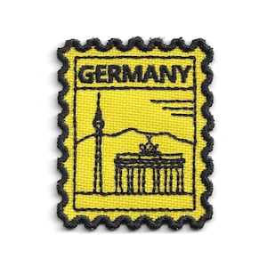 Può includere: Una toppa ricamata rettangolare a forma di francobollo con sfondo giallo e cuciture nere. Presenta la parola GERMANY in alto, con un contorno nero della torre della televisione di Berlino e della Porta di Brandeburgo sottostanti.