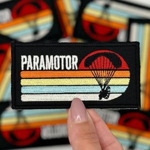 Può includere: Una toppa ricamata nera con un design di tramonto retrò e la parola "PARAMOTOR" in testo bianco. La toppa presenta un paracadute rosso e bianco con un motore attaccato.