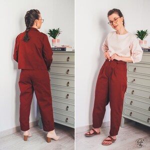 May include: A woman is wearing a maroon pantsuit with a cropped jacket and high-waisted pants. She is standing in front of a dresser with a book titled "The Moda" on top.