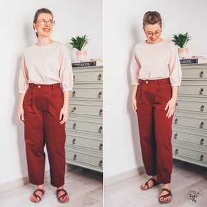 May include: A woman is wearing a light pink blouse with a slightly puffed sleeve and a pair of burgundy high-waisted pants. She is standing in front of a dresser with a book titled "EL MODA" on top.