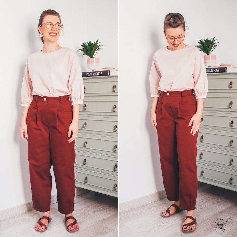May include: A woman is wearing a light pink blouse with a slightly puffed sleeve and a pair of burgundy high-waisted pants. She is standing in front of a dresser with a book titled "EL MODA" on top.