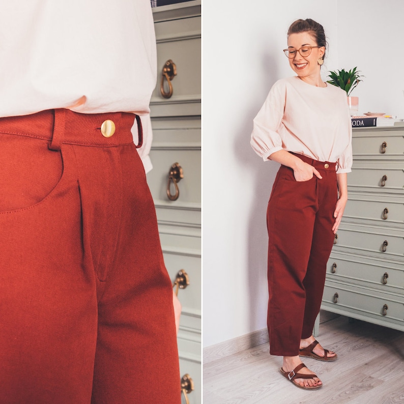 May include: A woman wearing a light pink blouse and rust-colored high-waisted pants with a gold button. The pants have a straight leg and are cropped above the ankle.
