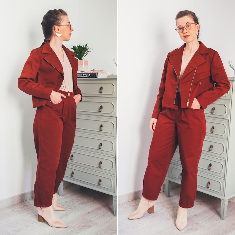 May include: Two women wearing matching red pantsuits with a blazer and pants. The pantsuits have a zipper closure and pockets. The women are wearing glasses and have their hair pulled back.