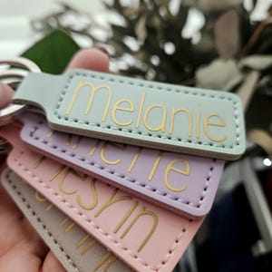 Könnte beinhalten: Eine Reihe personalisierter Schlüsselanhänger in Pastellfarben: Mintgrün, Lavendel, Rosa und Grau. Jeder Schlüsselanhänger zeigt einen Namen in goldfarbenen Buchstaben. Die Schlüsselanhänger sind rechteckig mit abgerundeten Ecken und genähten Kanten.