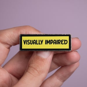 May include: A rectangular enamel pin featuring a bright yellow background with a thick black border. The text VISUALLY IMPAIRED is printed in bold black capital letters across the centre of the pin, which has a metallic silver-toned edge.