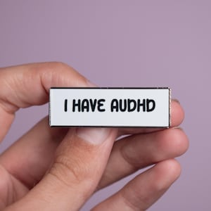 May include: A rectangular enamel pin with a white background and a thin metallic border. The text I HAVE AUDHD is printed in bold, black, rounded sans-serif capital letters across the centre of the pin, creating a clear and legible design.