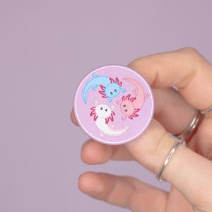 May include: A round, light purple button featuring three axolotl illustrations in blue, pink, and white. The axolotls are arranged in a circular pattern against a light purple background. The button is held by a hand.
