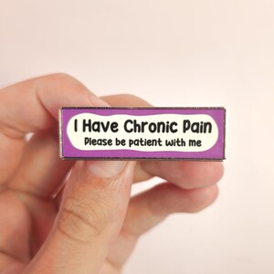 Puede incluir: Un pin rectangular con un borde morado y un centro blanco. El pin dice "I Have Chronic Pain" encima de "Please be patient with me" en texto negro. El pin está sostenido en la mano de una persona.