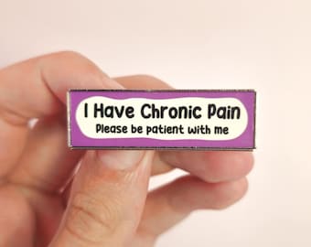May include: A rectangular pin with a purple border and a white centre. The pin reads "I Have Chronic Pain" above "Please be patient with me" in black text. The pin is held in a person's hand.
