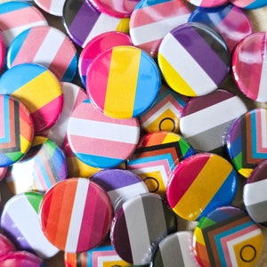 Puede incluir: Una colección de botones redondos coloridos con varios diseños de banderas del orgullo LGBTQ+. Los botones presentan rayas y patrones geométricos en tonos de rosa, azul, amarillo, morado y otros colores. Algunos están envueltos individualmente en plástico transparente.