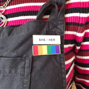 Könnte beinhalten: Schwarze Canvas-Schürze mit zwei Anstecknadeln. Die obere Anstecknadel trägt die Aufschrift "SHE / HER" in weißer Schrift auf goldenem Hintergrund. Die untere Anstecknadel ist ein Regenbogenflaggen-Design mit roten, orangen, gelben, grünen, blauen und violetten Streifen.