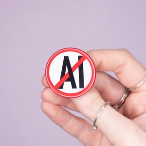 May include: A circular badge with a red border, featuring the letters "AI" crossed out with a red diagonal line. The badge is held in a hand with silver rings, set against a lilac backdrop.
