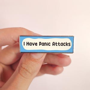 Peut inclure: Une épingle rectangulaire en émail avec le texte "I Have Panic Attacks" en lettres noires sur fond blanc, encadrée d'une bordure bleu clair. L'épingle est tenue par une main sur un fond neutre et doux.