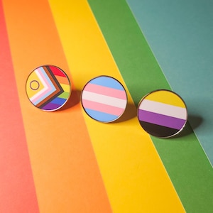 May include: Three circular enamel pins with silver edges. The pins display the Progress Pride flag, the transgender flag, and the non-binary flag. The background is a rainbow of colours.