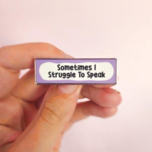 May include: A rectangular enamel pin with a silver border. The pin features the text "Sometimes I Struggle To Speak" in black font on a white background, framed by a light purple border. The pin is held in a hand against a soft pink background.