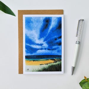 May include: A watercolour painting of a beach scene with a dramatic blue sky, ocean, and sandy shore. The artwork is on a white card, inside a brown envelope. A white pen with a crystal-like design is next to the card.