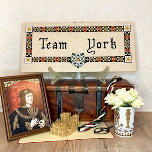 Puede incluir: Una colección de artículos que incluyen un retrato enmarcado, un letrero bordado a punto de cruz que dice "Team York", un cofre de madera, una taza y una maqueta de papel de un edificio. El letrero tiene un borde floral y un emblema de rosa blanca.