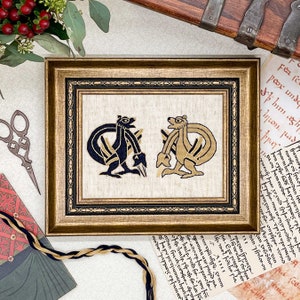 May include: A framed cross-stitch embroidery featuring two stylized animal figures, one in black and one in gold, against a cream-colored background. The frame is gold with a black inner border.