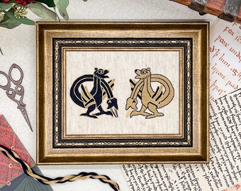 May include: A framed cross-stitch embroidery featuring two stylized animal figures, one in black and one in gold, against a cream-colored background. The frame is gold with a black inner border.