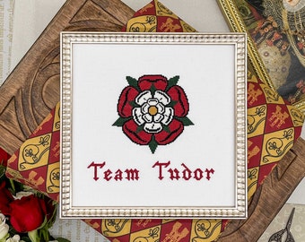 May include: Cross-stitch framed art featuring the Tudor rose emblem in red, white, and green, with the words "Team Tudor" stitched in red. The artwork is displayed on a patterned fabric with a wooden frame.