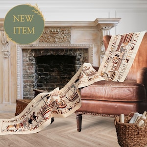 May include: A long, cream-colored fabric runner featuring a detailed historical narrative print in black, red, and brown. The design depicts stylized ships, figures, and horses in a continuous sequence, draped over a brown leather armchair.