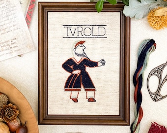 May include: A framed cross-stitch embroidery featuring a stylized figure with the name "TVROLD" above it. The figure is wearing a blue and red outfit and is standing with one hand on its hip.