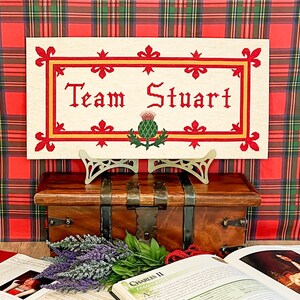 Peut inclure: Un sampler de point de croix blanc avec une bordure rouge et or. Le sampler présente le texte "Team Stuart" et un chardon stylisé.