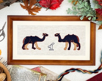 May include: A framed cross-stitch embroidery featuring two black camels with brown accents on a beige background. The camels are facing opposite directions and are surrounded by a white border.